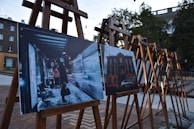 Several easels are set up outdoors, each displaying photographs. The image in the foreground shows a nighttime scene at a bus stop with a person in a long coat and a bright vest, holding a bag. The second image depicts a brick building with a car parked in front.