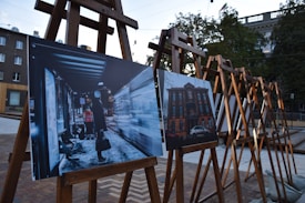 Several easels are set up outdoors, each displaying photographs. The image in the foreground shows a nighttime scene at a bus stop with a person in a long coat and a bright vest, holding a bag. The second image depicts a brick building with a car parked in front.