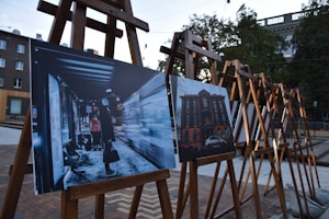 Several easels are set up outdoors, each displaying photographs. The image in the foreground shows a nighttime scene at a bus stop with a person in a long coat and a bright vest, holding a bag. The second image depicts a brick building with a car parked in front.