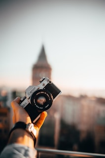 A compact digital camera held in a hand against a blurred cityscape background.