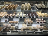 A bakery display case filled with an assortment of cupcakes and cookies. The cupcakes vary in flavors and toppings, including chocolate, vanilla, and sprinkled varieties, arranged in neat rows on multiple shelves. Signage highlights the names and prices of the baked goods. The overall presentation is colorful and appetizing.