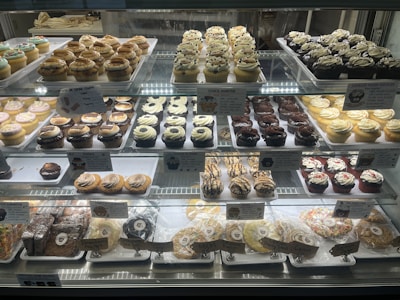 A bakery display case filled with an assortment of cupcakes and cookies. The cupcakes vary in flavors and toppings, including chocolate, vanilla, and sprinkled varieties, arranged in neat rows on multiple shelves. Signage highlights the names and prices of the baked goods. The overall presentation is colorful and appetizing.