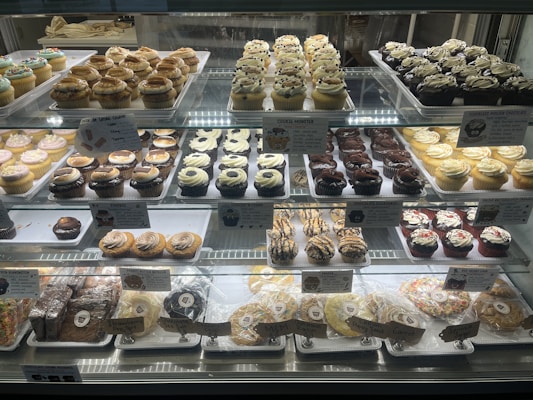 A bakery display case filled with an assortment of cupcakes and cookies. The cupcakes vary in flavors and toppings, including chocolate, vanilla, and sprinkled varieties, arranged in neat rows on multiple shelves. Signage highlights the names and prices of the baked goods. The overall presentation is colorful and appetizing.
