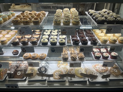 A bakery display case filled with an assortment of cupcakes and cookies. The cupcakes vary in flavors and toppings, including chocolate, vanilla, and sprinkled varieties, arranged in neat rows on multiple shelves. Signage highlights the names and prices of the baked goods. The overall presentation is colorful and appetizing.