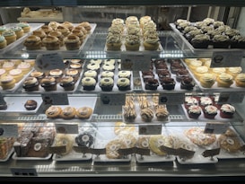 A bakery display case filled with an assortment of cupcakes and cookies. The cupcakes vary in flavors and toppings, including chocolate, vanilla, and sprinkled varieties, arranged in neat rows on multiple shelves. Signage highlights the names and prices of the baked goods. The overall presentation is colorful and appetizing.