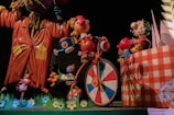 A whimsical scene featuring colorful, cartoon-like characters. A large, animated figure in an orange coat stands on the left, holding a bird. A cheerful character with a red nose is riding a bicycle with a wheel in a multicolored pattern. Other smaller characters appear to be interacting playfully around them. The setting includes vibrant, oversized flowers and a checkered wall.