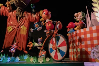 A whimsical scene featuring colorful, cartoon-like characters. A large, animated figure in an orange coat stands on the left, holding a bird. A cheerful character with a red nose is riding a bicycle with a wheel in a multicolored pattern. Other smaller characters appear to be interacting playfully around them. The setting includes vibrant, oversized flowers and a checkered wall.