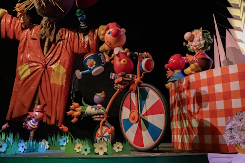A whimsical scene featuring colorful, cartoon-like characters. A large, animated figure in an orange coat stands on the left, holding a bird. A cheerful character with a red nose is riding a bicycle with a wheel in a multicolored pattern. Other smaller characters appear to be interacting playfully around them. The setting includes vibrant, oversized flowers and a checkered wall.