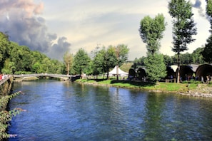 A serene river flows gently alongside a lush green park with tall trees and neatly maintained grassy areas. In the background, a bridge spans the river, connecting both sides of the park. People are seen strolling and gathering, suggesting a lively yet peaceful environment. The sky above is partly cloudy, with hints of sunlight breaking through.