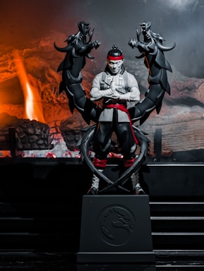 A stylized statue of a martial artist stands amidst a dramatic fiery backdrop. The character is clad in a white robe with a red headband and belt. Black ornamental dragons coil around him, enhancing the intense and mystical atmosphere.