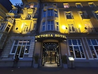An elegant hotel facade with classical architectural details, warmly illuminated by exterior lights at dusk. The entrance is centrally located, featuring a curved awning with the hotel's name in illuminated letters. Tall windows are symmetrically arranged on the building's facade.