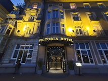 An elegant hotel facade with classical architectural details, warmly illuminated by exterior lights at dusk. The entrance is centrally located, featuring a curved awning with the hotel's name in illuminated letters. Tall windows are symmetrically arranged on the building's facade.
