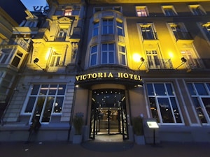 An elegant hotel facade with classical architectural details, warmly illuminated by exterior lights at dusk. The entrance is centrally located, featuring a curved awning with the hotel's name in illuminated letters. Tall windows are symmetrically arranged on the building's facade.