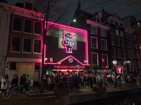 A lively urban night scene featuring a building decorated with bright neon lights, including a prominently displayed pink and blue neon artwork of a figure. Crowds of people are gathered outside, suggesting a popular attraction or event. Bicycles are visible along the street, adding to the bustling city atmosphere. The architecture includes classic multi-story brick buildings.