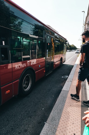 A red city bus is parked on the street with the words 'ROMA atac' visible on its side. A person is standing nearby on the sidewalk, partially visible in the right corner, with a green shopping bag.