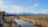 A photographer is capturing images of moose walking through a shallow stream in a vast, open landscape. Snow-capped mountains rise in the background under a clear blue sky, while dry grasses line the sides of the stream.