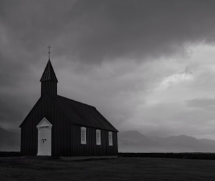 A solitary black wooden church with white trim stands under a dramatic, overcast sky. The building features a tall, pointed steeple with a cross at the peak. Surrounding the church is an open field, with distant, shadowy mountains in the background suggesting a remote location.