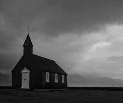 A solitary black wooden church with white trim stands under a dramatic, overcast sky. The building features a tall, pointed steeple with a cross at the peak. Surrounding the church is an open field, with distant, shadowy mountains in the background suggesting a remote location.