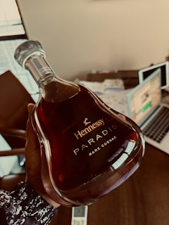 A person is holding a bottle of Hennessy Paradis Rare Cognac, which has an elegant, curved design and contains a deep amber liquid inside. The background includes a desk with papers, a laptop, and a chair, suggesting an office or study environment.