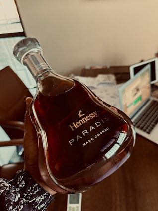 A person is holding a bottle of Hennessy Paradis Rare Cognac, which has an elegant, curved design and contains a deep amber liquid inside. The background includes a desk with papers, a laptop, and a chair, suggesting an office or study environment.