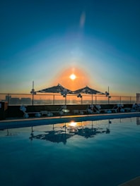 A serene rooftop swimming pool reflecting the vibrant hues of a sunset, with umbrellas and lounge chairs neatly arranged along the edge. The sun casts a warm glow across the sky, creating a peaceful and tranquil atmosphere.