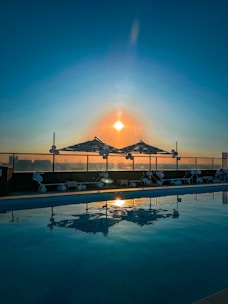 A serene rooftop swimming pool reflecting the vibrant hues of a sunset, with umbrellas and lounge chairs neatly arranged along the edge. The sun casts a warm glow across the sky, creating a peaceful and tranquil atmosphere.