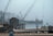 A foggy waterfront scene with a wooden dock in the foreground, covered with lobster traps and fishing equipment. A small, rustic wooden shed stands on the dock. In the background, a large crane on a barge with the word 'Winslow' is visible through the mist. The water is calm, and a small boat is moored nearby.