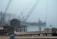 A foggy waterfront scene with a wooden dock in the foreground, covered with lobster traps and fishing equipment. A small, rustic wooden shed stands on the dock. In the background, a large crane on a barge with the word 'Winslow' is visible through the mist. The water is calm, and a small boat is moored nearby.