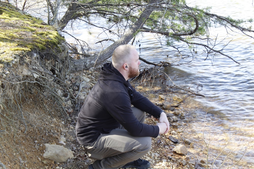 a man squatting on a rocky shore next to a body of water