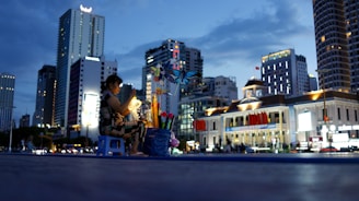 A woman sits on a small blue stool, tending to a stall decorated with vibrant pinwheels and lit by a warm lamp. The scene unfolds against a backdrop of tall, illuminated buildings and a historic-looking structure adorned with red flags, under the dusky sky.