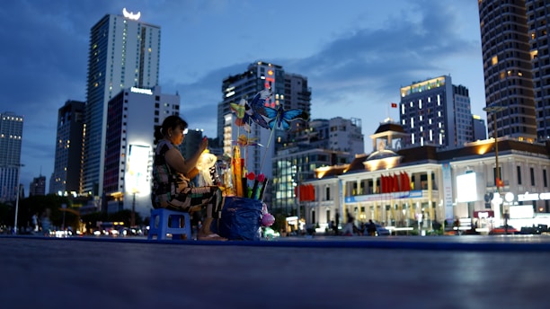 A woman sits on a small blue stool, tending to a stall decorated with vibrant pinwheels and lit by a warm lamp. The scene unfolds against a backdrop of tall, illuminated buildings and a historic-looking structure adorned with red flags, under the dusky sky.