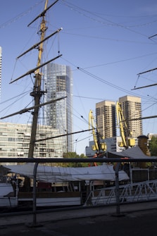 A tall ship with intricate rigging is docked at a harbor, set against a backdrop of modern high-rise buildings. The ship's mast and sails are prominent, while cranes and other industrial equipment are visible nearby. The scene combines elements of maritime history with contemporary urban architecture under a clear blue sky.