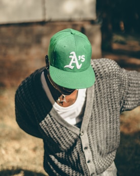 A person wearing a green cap with a white 'A's' logo, a gray knit cardigan, and a white shirt is seen bending forward outdoors. The background has a blurred mix of earthy tones and a structure that might be a wall.