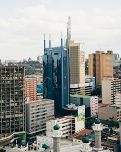 A cityscape featuring tall office buildings with diverse architectural designs. Prominent among the structures is a blue glass building with vertical accents and a tan building with a large advertisement. The foreground includes a mosque with green domes, contrasting modern and traditional architecture.