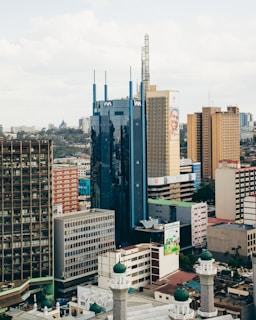 A cityscape featuring tall office buildings with diverse architectural designs. Prominent among the structures is a blue glass building with vertical accents and a tan building with a large advertisement. The foreground includes a mosque with green domes, contrasting modern and traditional architecture.