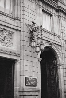 A grand architectural facade featuring intricate stonework and ornate details, including sculpted figures and decorative iron lamps. The entrance is marked with a sign for the Museo Nacional de Arte.