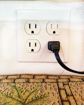A wall electrical outlet with four sockets, with one socket currently in use by a black power plug. The outlet is mounted on a white textured wall, and in front of it is a textured surface with a pattern resembling a woven fabric or decorative mat.