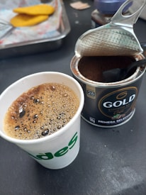 A paper cup filled with freshly brewed coffee sits next to an open can of ground coffee with a spoon inside. The coffee in the cup is frothy and the can has a dark label. In the background, there is a tray with pieces of bread.