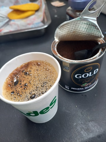 A paper cup filled with freshly brewed coffee sits next to an open can of ground coffee with a spoon inside. The coffee in the cup is frothy and the can has a dark label. In the background, there is a tray with pieces of bread.