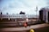 A railway scene featuring a train with multiple carriages parked at the station. A worker in a safety vest and helmet is walking across the platform holding a red flag. Another worker is visible near the train. The sky is cloudy and there are trees in the background.