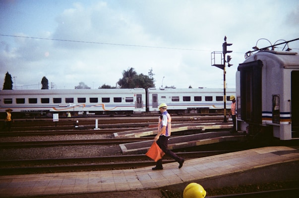 A railway scene featuring a train with multiple carriages parked at the station. A worker in a safety vest and helmet is walking across the platform holding a red flag. Another worker is visible near the train. The sky is cloudy and there are trees in the background.