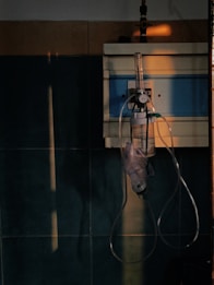 A dimly lit hospital room featuring an oxygen regulator attached to a wall. The device includes a flow meter and oxygen mask with tubing. The walls are tiled, with some areas catching a warm light.