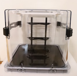 A transparent cube-shaped container with black latches and a sleek black interior stand or support structure. The container is made of clear plastic or glass, and the black components provide a contrast.