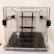 A transparent cube-shaped container with black latches and a sleek black interior stand or support structure. The container is made of clear plastic or glass, and the black components provide a contrast.