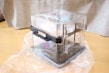 A transparent plastic storage container with black handles resting on a wooden floor. The container is partially wrapped in plastic. The background includes a gray fabric, possibly a curtain or a piece of furniture cover.