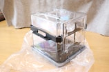 A transparent plastic storage container with black handles resting on a wooden floor. The container is partially wrapped in plastic. The background includes a gray fabric, possibly a curtain or a piece of furniture cover.