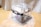 A transparent plastic storage container with black handles resting on a wooden floor. The container is partially wrapped in plastic. The background includes a gray fabric, possibly a curtain or a piece of furniture cover.