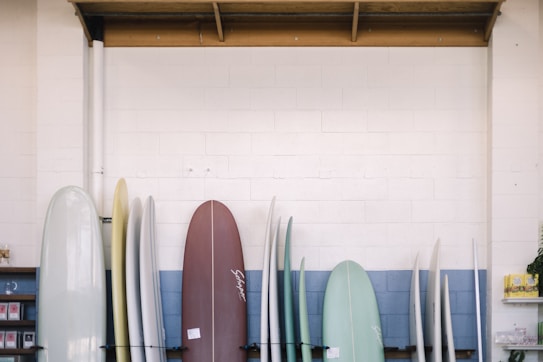 A collection of surfboards is lined up against a wall inside a shop. The wall features a blue and white color scheme, and there is shelving on the left holding various small items. A wooden beam runs along the ceiling above the surfboards. The surfboards vary in color, including white, cream, brown, and soft green.