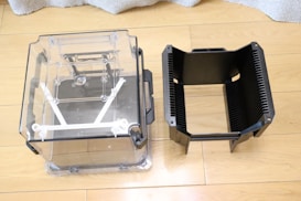 Two plastic containers are placed side by side on a wooden floor. The one on the left is transparent with clear sides showing its internal structure, and the one on the right has black, vertical slats and a more solid appearance. Both have handles on their sides.