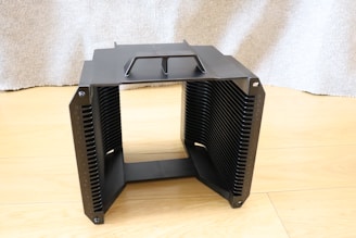 A black, rectangular heat sink with multiple fins is placed on a light wooden floor. The top part has a handle-like structure. The background is a textured grey fabric or curtain.